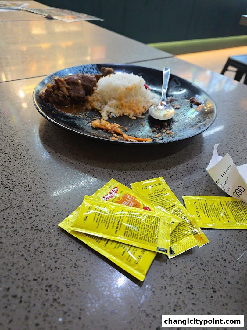 A plate of rice and meat with chili sauce packets on a table.