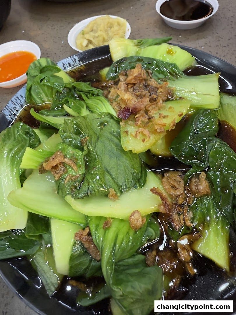 A plate of stir-fried bok choy with crispy fried shallots and a side of chili sauce.