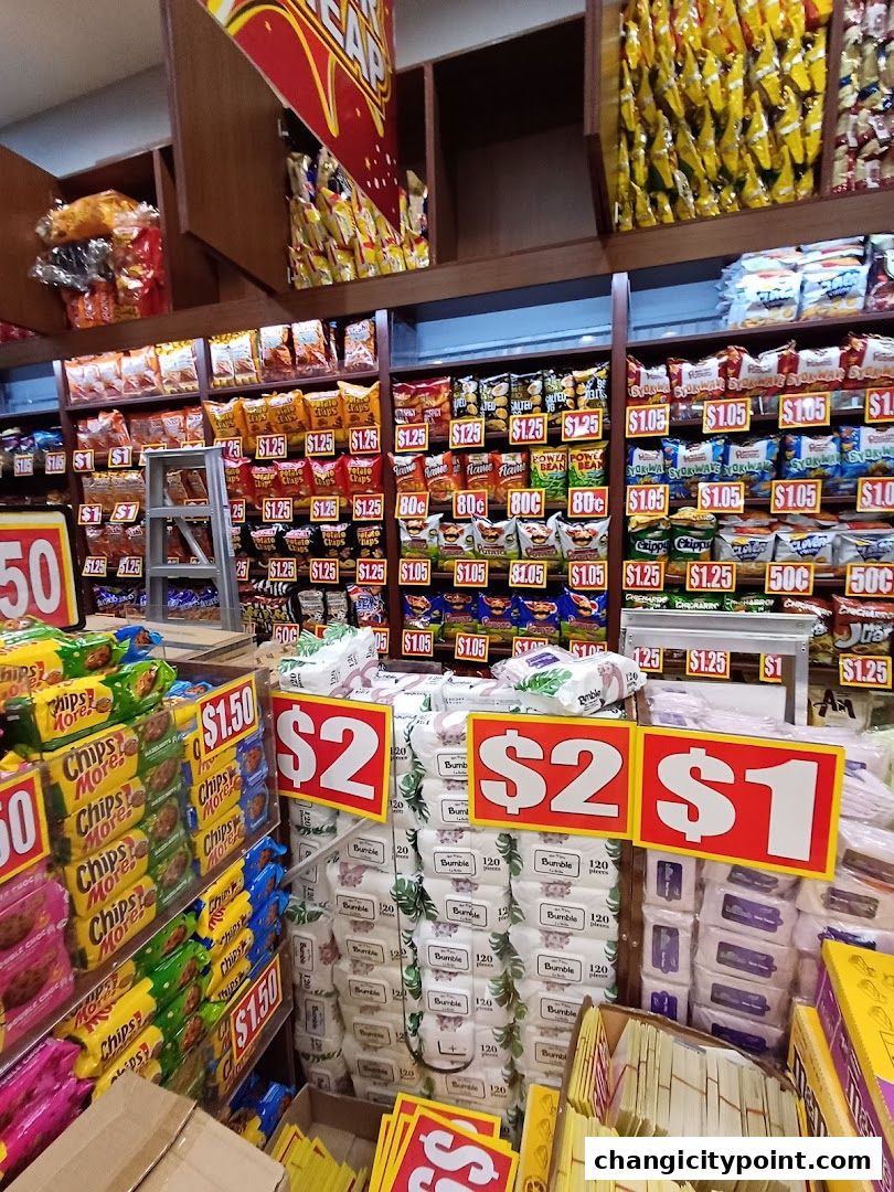 A retail shop interior displaying shelves stocked with various snacks and household items, with prominent price signage.