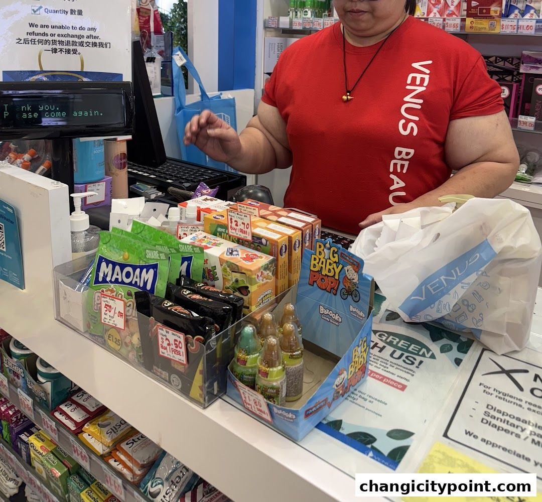 A cashier in a red Venus Beauty t-shirt stands behind a counter with various products and bags.