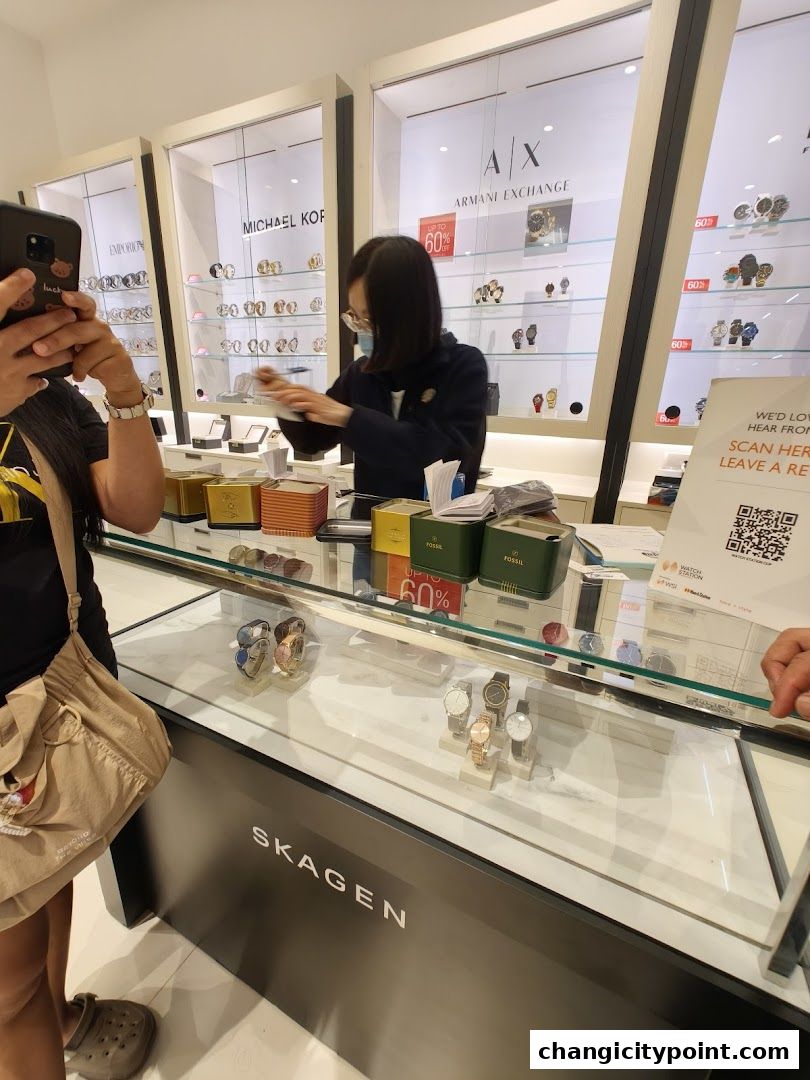 A store employee assists a customer at a watch counter displaying various brands.