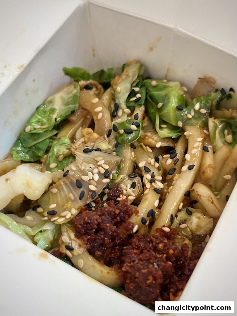 A close-up of a takeaway box filled with noodles, vegetables, and chili paste.
