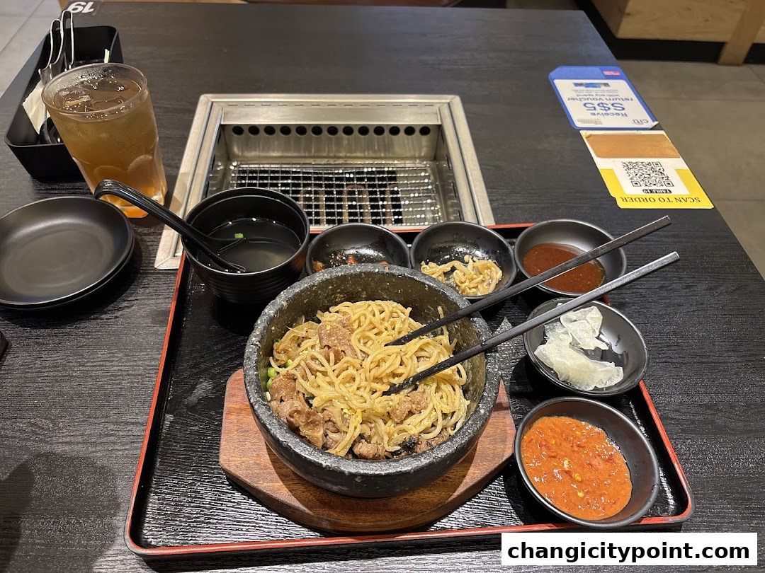 A stone bowl of noodles with meat and side dishes on a table.